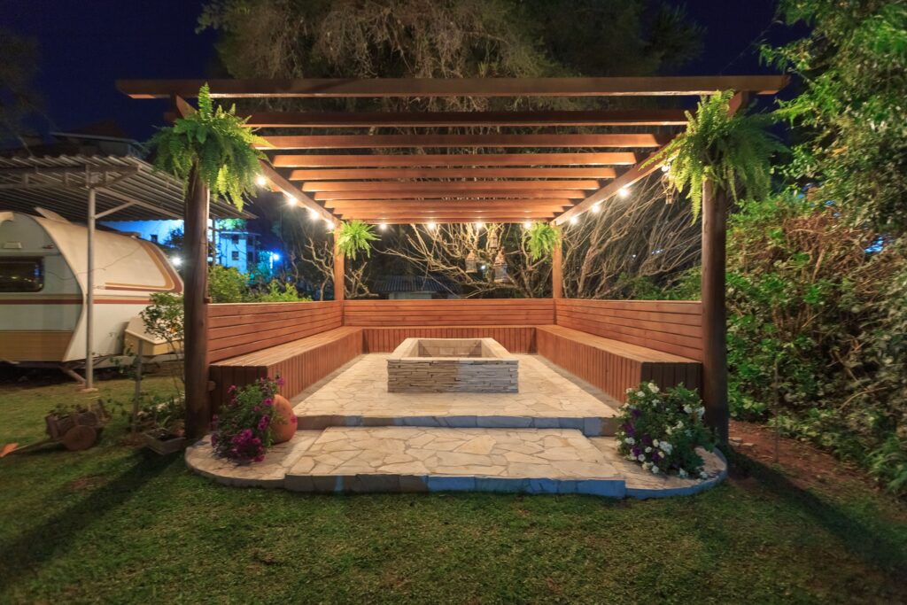 Illuminated wooden gazebo with fireplace and benches in garden setting at night.